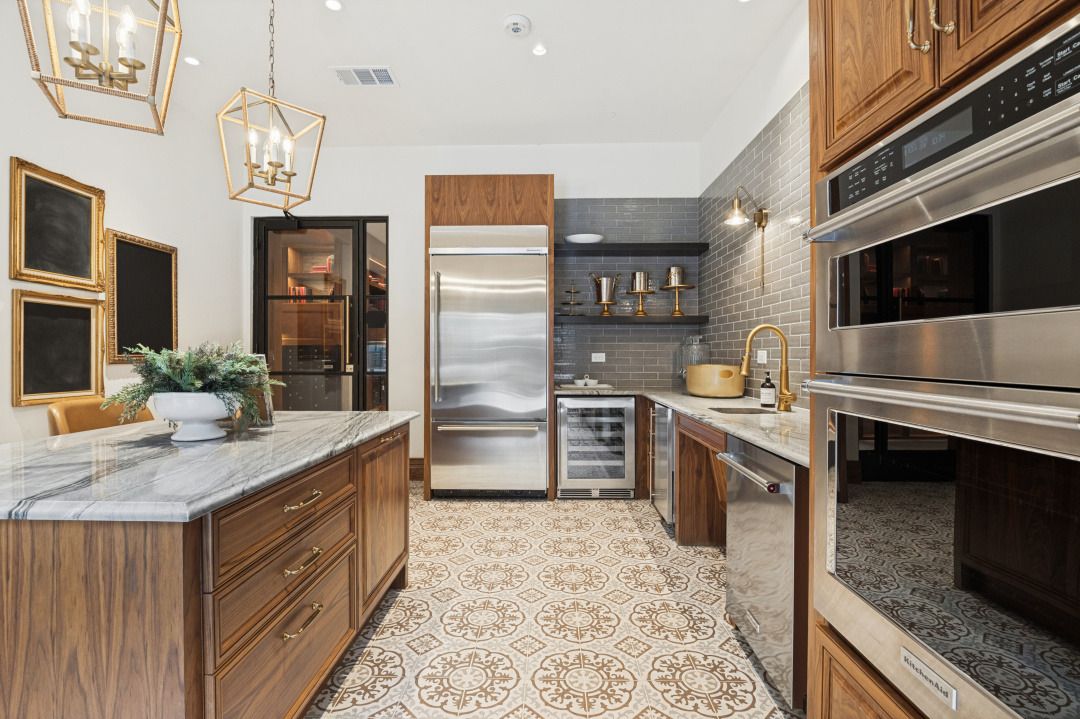 A modern kitchen with wood cabinets, a marble-topped island, stainless steel appliances, and patterned tile flooring.