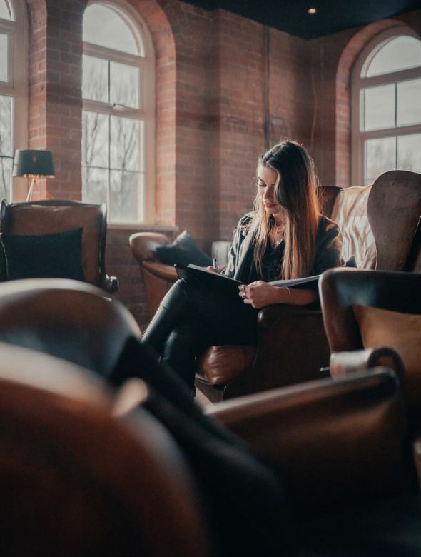 Woman sitting in a leather chair by large windows, writing in a notebook in a cozy, softly lit room.