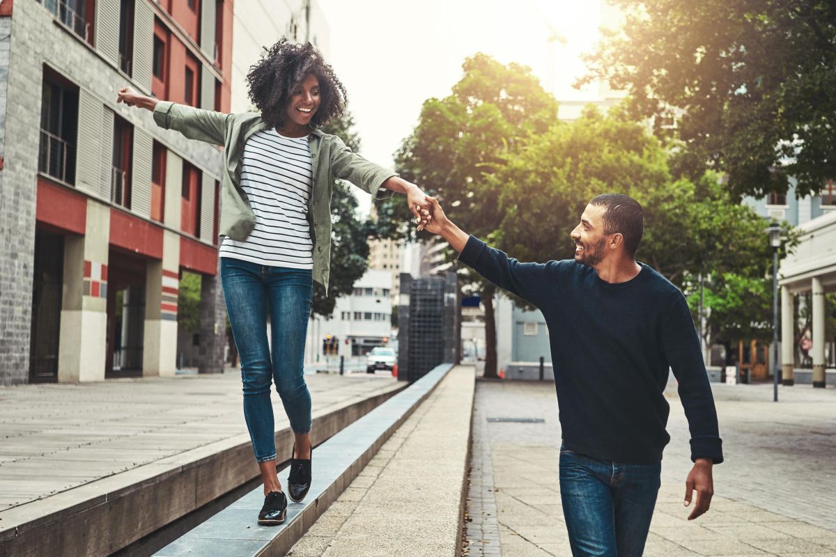A smiling woman balances on a ledge, holding hands with a man walking beside her in a city street.